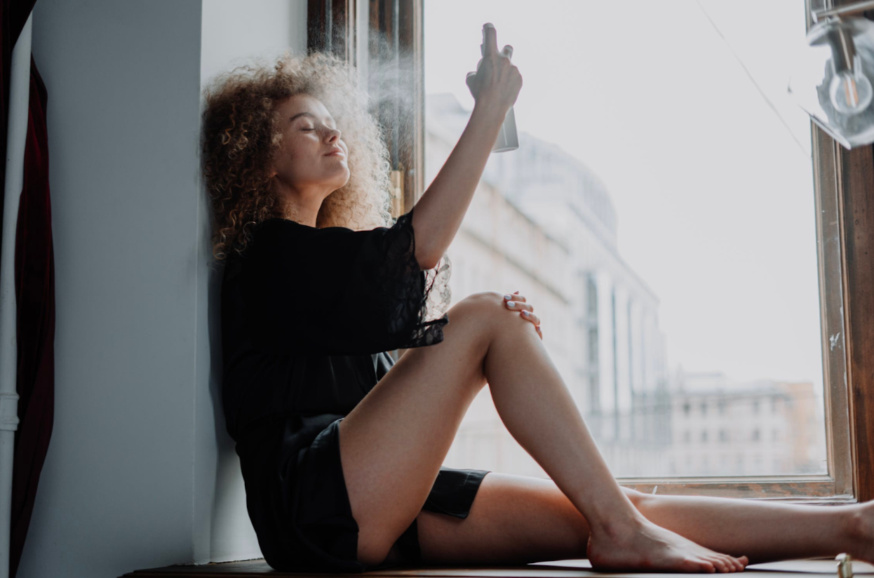 woman sitting in front of a window misting herself with sunscreen spray