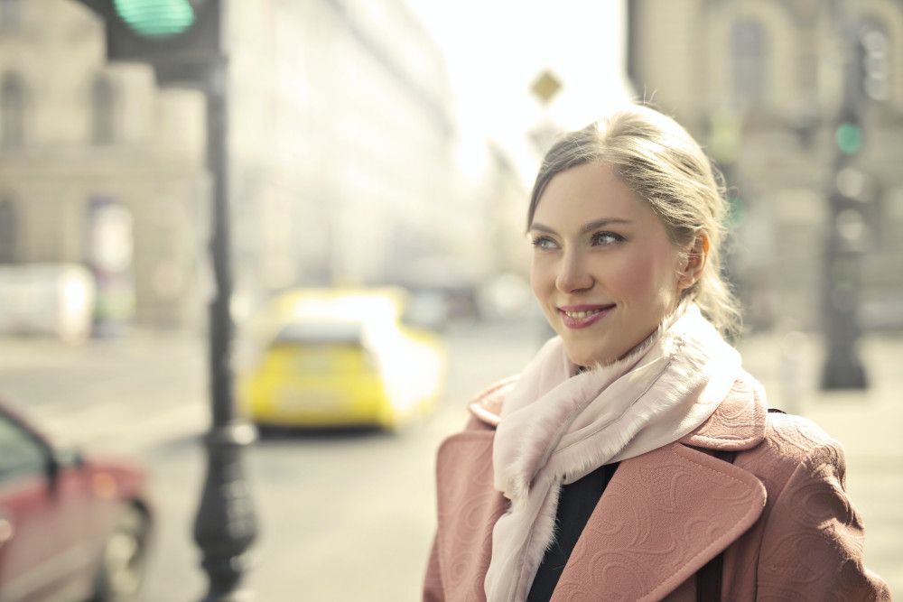 A woman wearing a pink coat and scarf stands outdoors in an urban setting, with cars and buildings in the background.