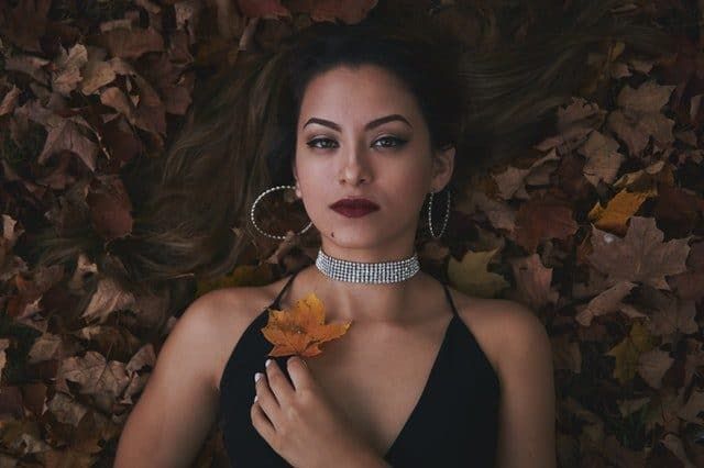 A woman with long hair, large hoop earrings, and a rhinestone choker lies on brown autumn leaves, holding a single leaf to her chest.