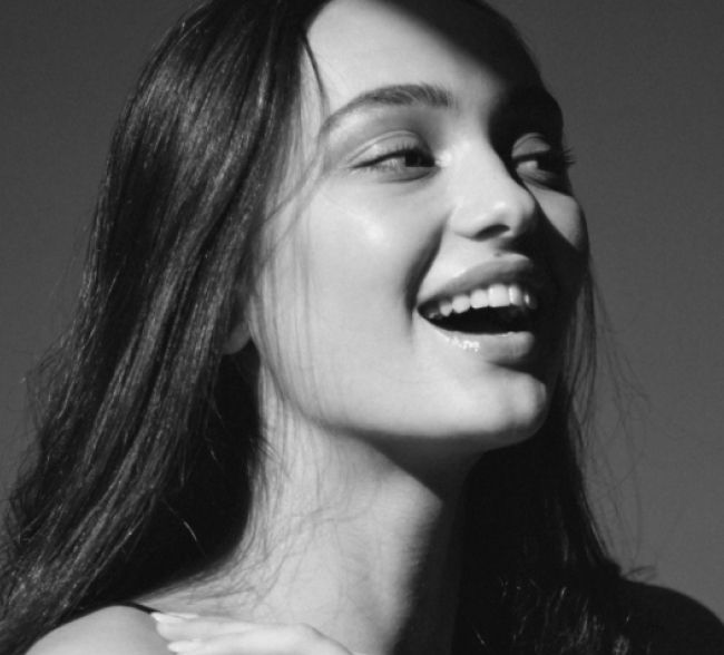 Black and white photo of a young woman with long hair smiling and looking slightly upward, with a shadow partially covering her forehead.