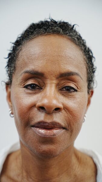 A close-up of an older woman with short, curly hair, wearing small hoop earrings and a white top, facing the camera with a neutral expression.
