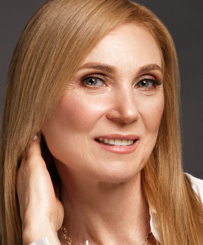 A woman with straight, light brown hair smiles slightly, touching her neck, with a neutral gray background.
