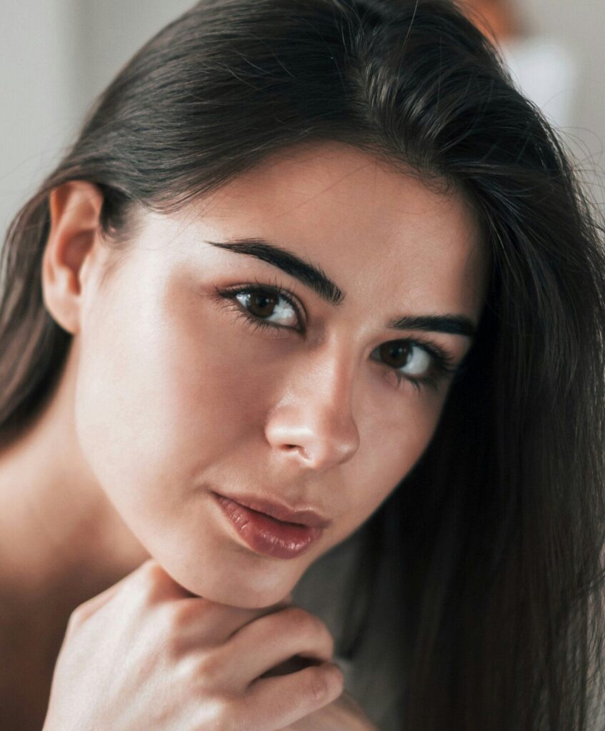 A woman with long dark hair looks directly at the camera, resting her chin on her hand. She has natural makeup and a neutral expression.