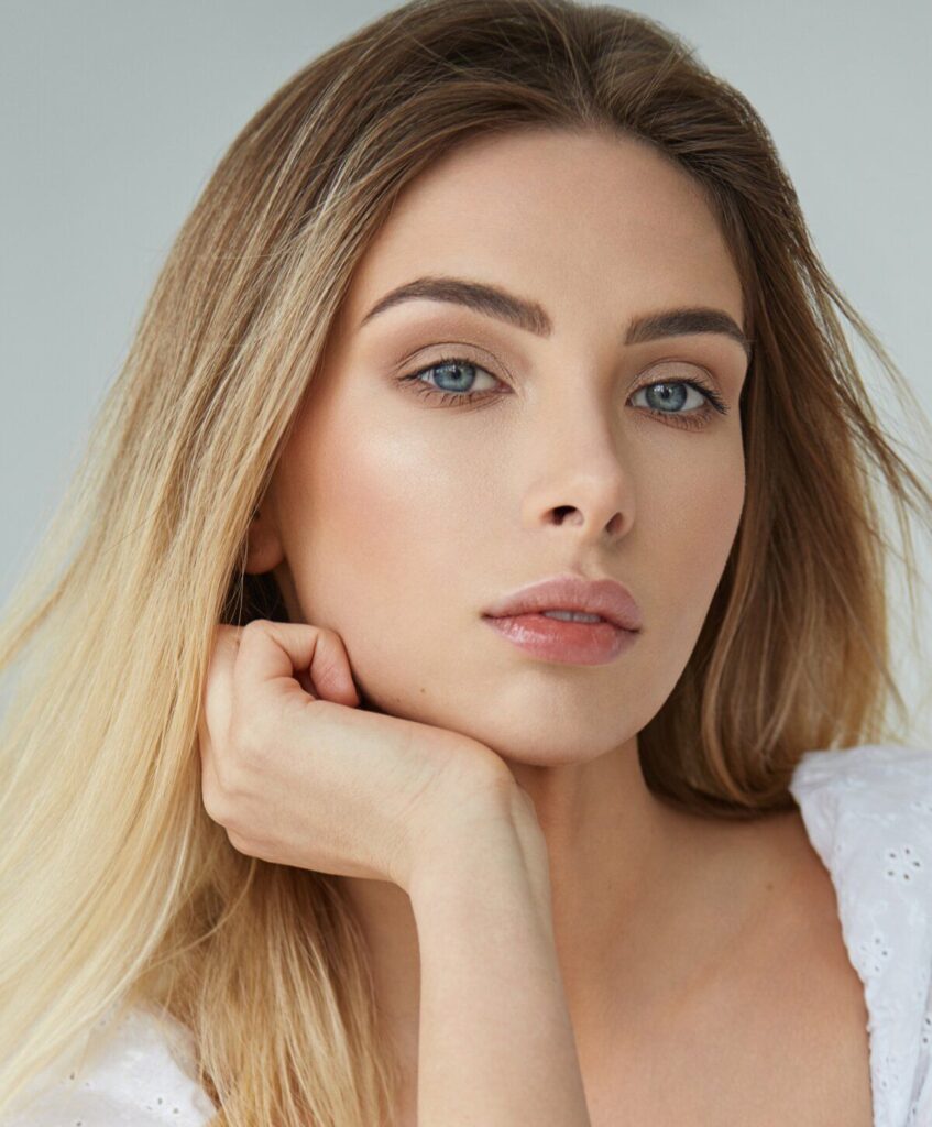 Woman with long blonde hair, neutral makeup, and blue eyes rests her chin on her hand, looking directly at the camera.